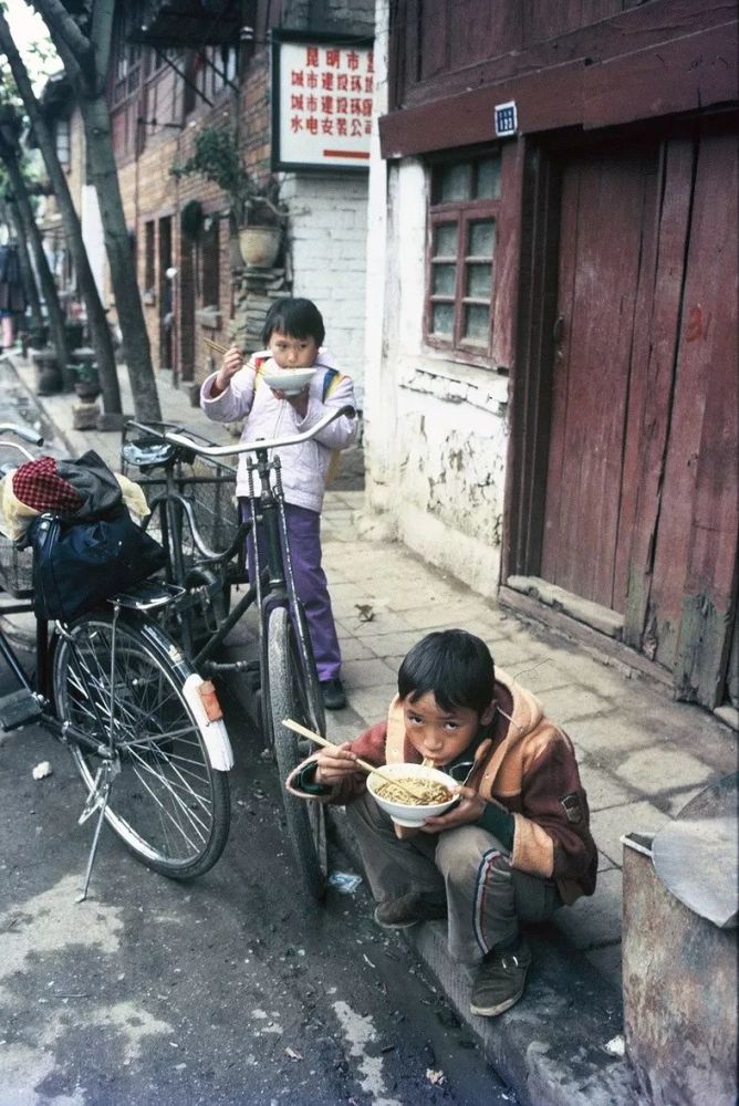 1985年,昆明,在一条富有年代感的街道边,有两个小孩在吃面.