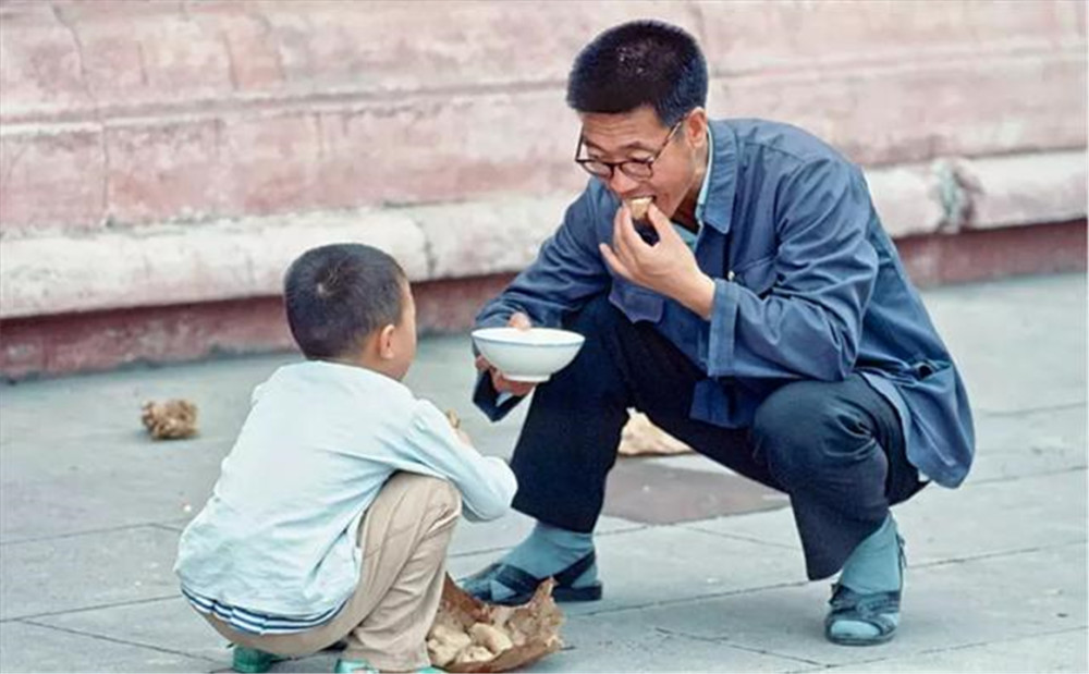 一个父亲和儿子蹲在地上吃包子,父亲手上还拿着一碗水.