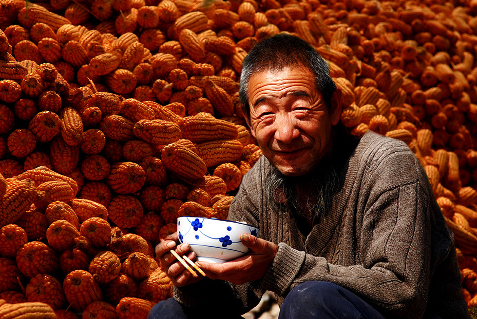 这是一幅拍摄于山区的老农民形象,坐在丰收的玉米前,从他的笑容里