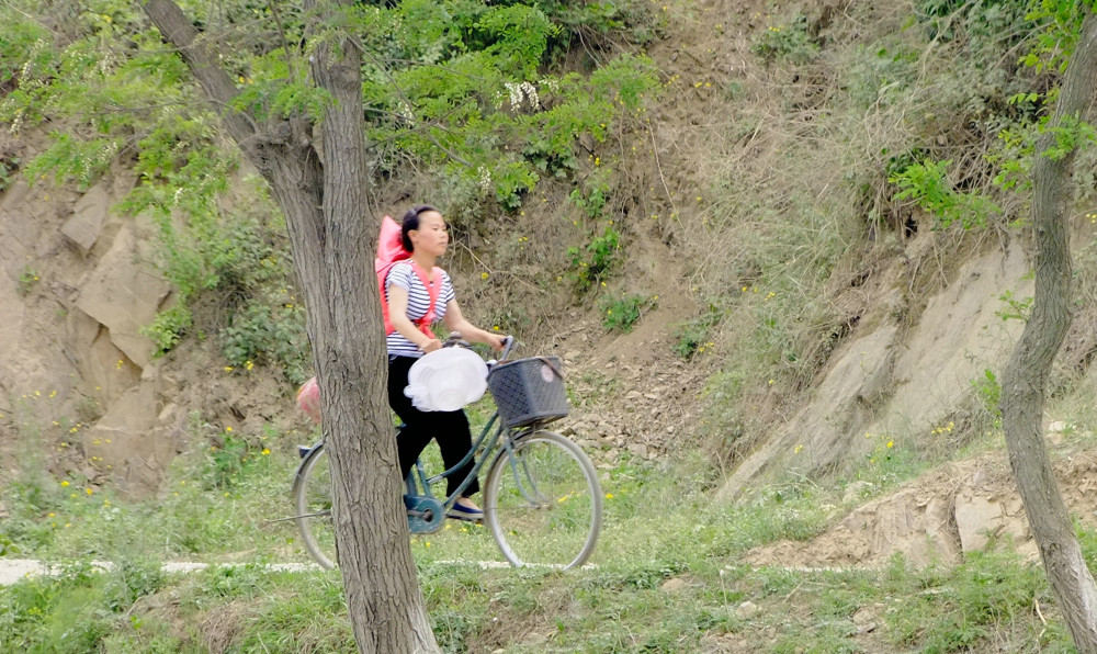 图为朝鲜农村的山路上,一名朝鲜女性背着小孩,骑着自行车.