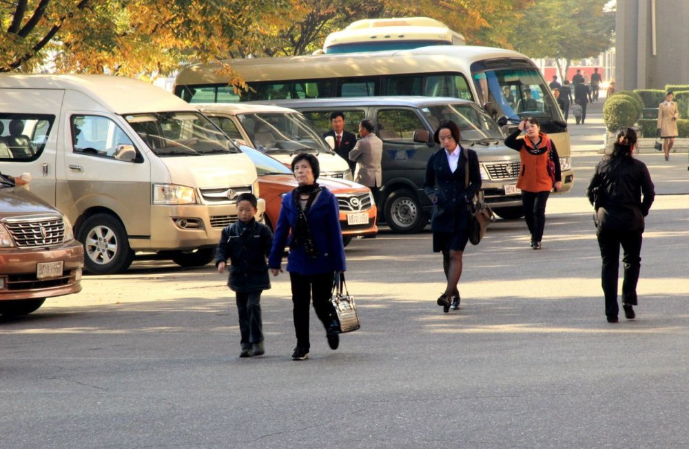 图为朝鲜街头步行的路人,一名女子穿着黑丝袜,旁边停着很多汽车.