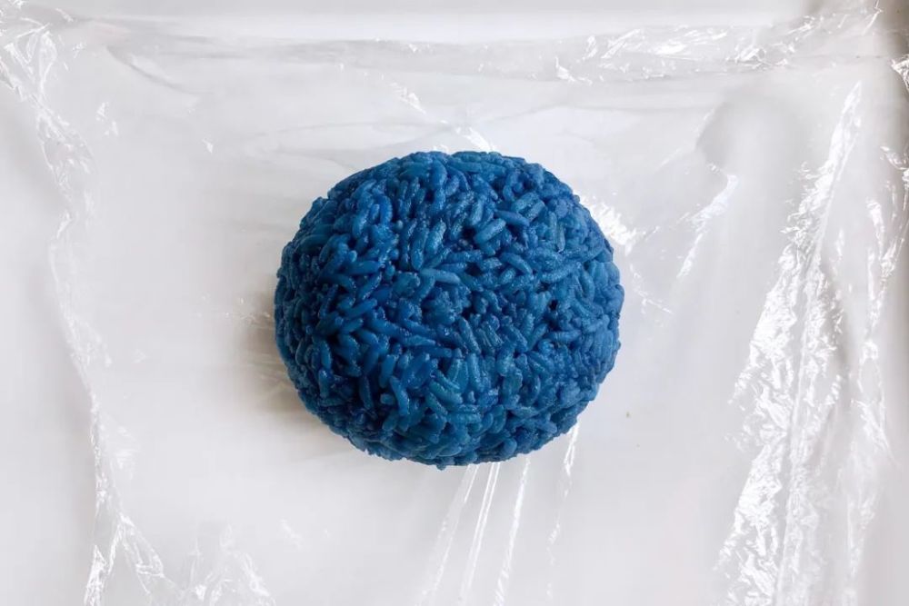 要尽量捏紧些才不容易散6,盘子上垫张保鲜膜,铺上一碗煮好的蓝色米饭