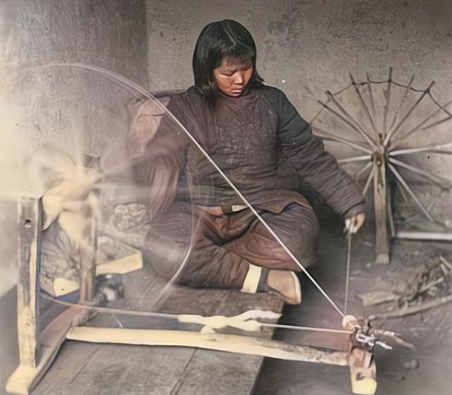 纺线的女子盘腿而坐,操作传统的防线工具,一手摇着大的滚轮,一手捏着