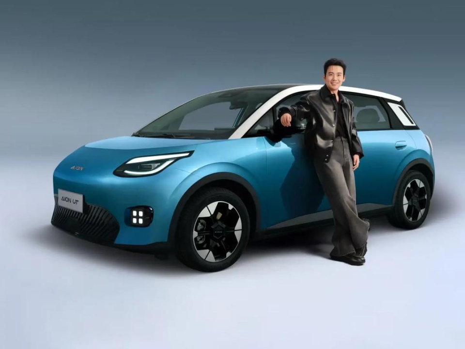 A young man in a black jacket and gray pants is leaning against a blue AION U T super electric vehicle, showcasing its modern design and branding.