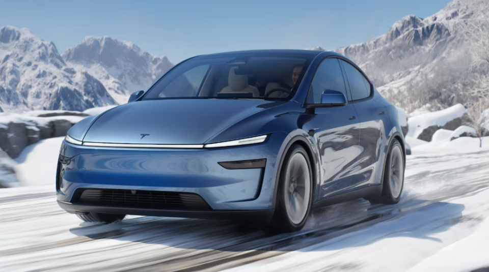 A sleek blue Tesla Model 3 is driving on a snow-covered mountain road, showcasing its streamlined design amid a stunning winter landscape with snow-capped peaks in the background.